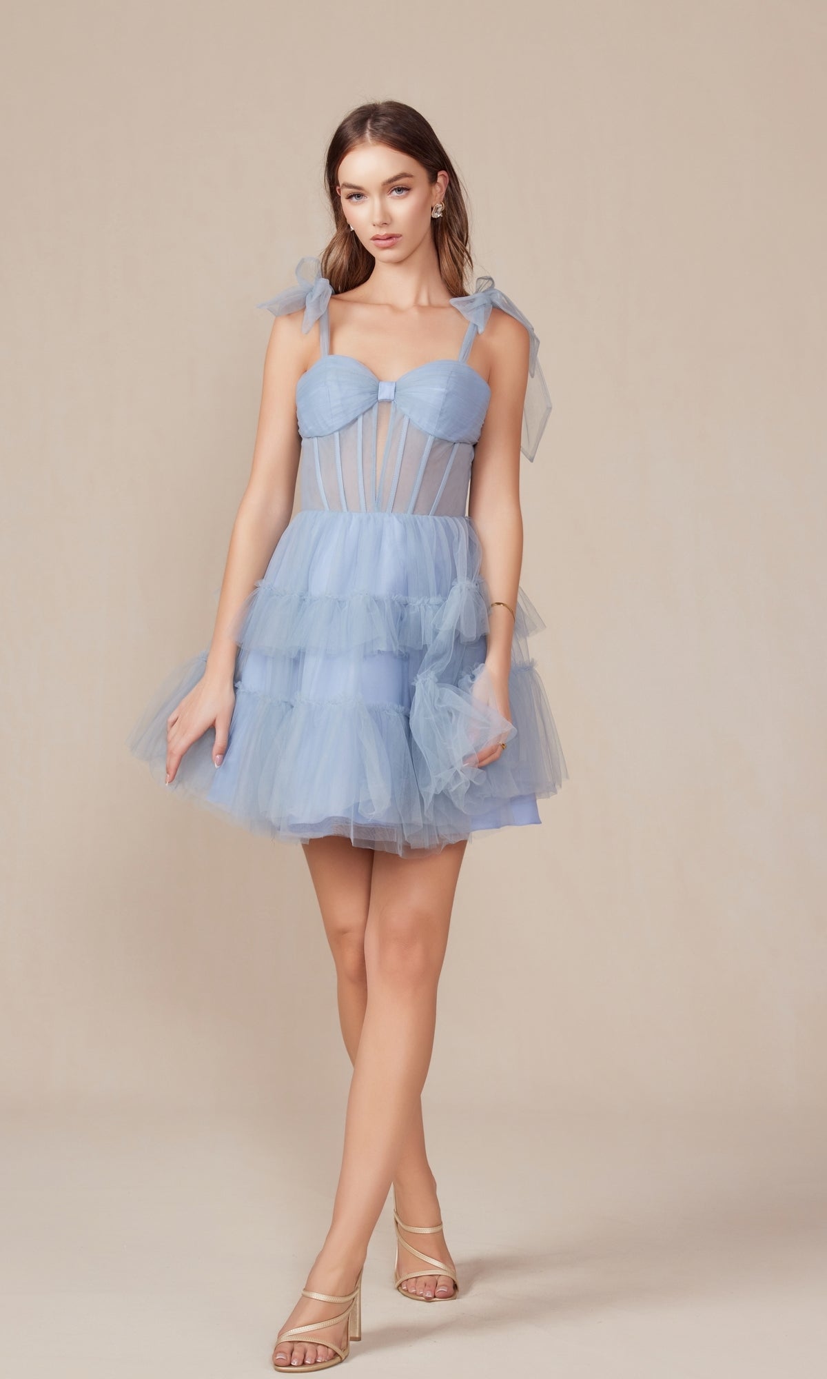 A woman wears the Shoulder-Tie Short Ruffled A-Line Hoco Dress Y858 in light blue tulle with sheer panels, a sheer corset bodice, and tiered ruffles, paired with nude heeled sandals against a plain beige background.